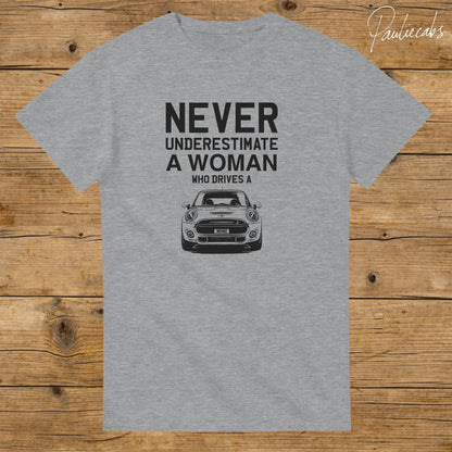 May include: A heather gray short-sleeved t-shirt featuring black text that reads NEVER UNDERESTIMATE A WOMAN WHO DRIVES A, positioned above a black and white graphic illustration of a Mini Cooper car with the word MINI on the license plate.