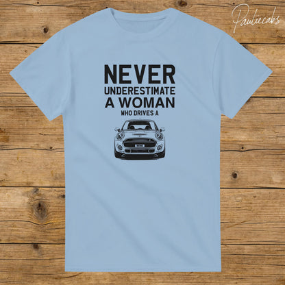 May include: A light blue short-sleeved t-shirt featuring black text that reads NEVER UNDERESTIMATE A WOMAN WHO DRIVES A, positioned above a black and white graphic illustration of the front view of a Mini Cooper car with the word MINI on the plate.