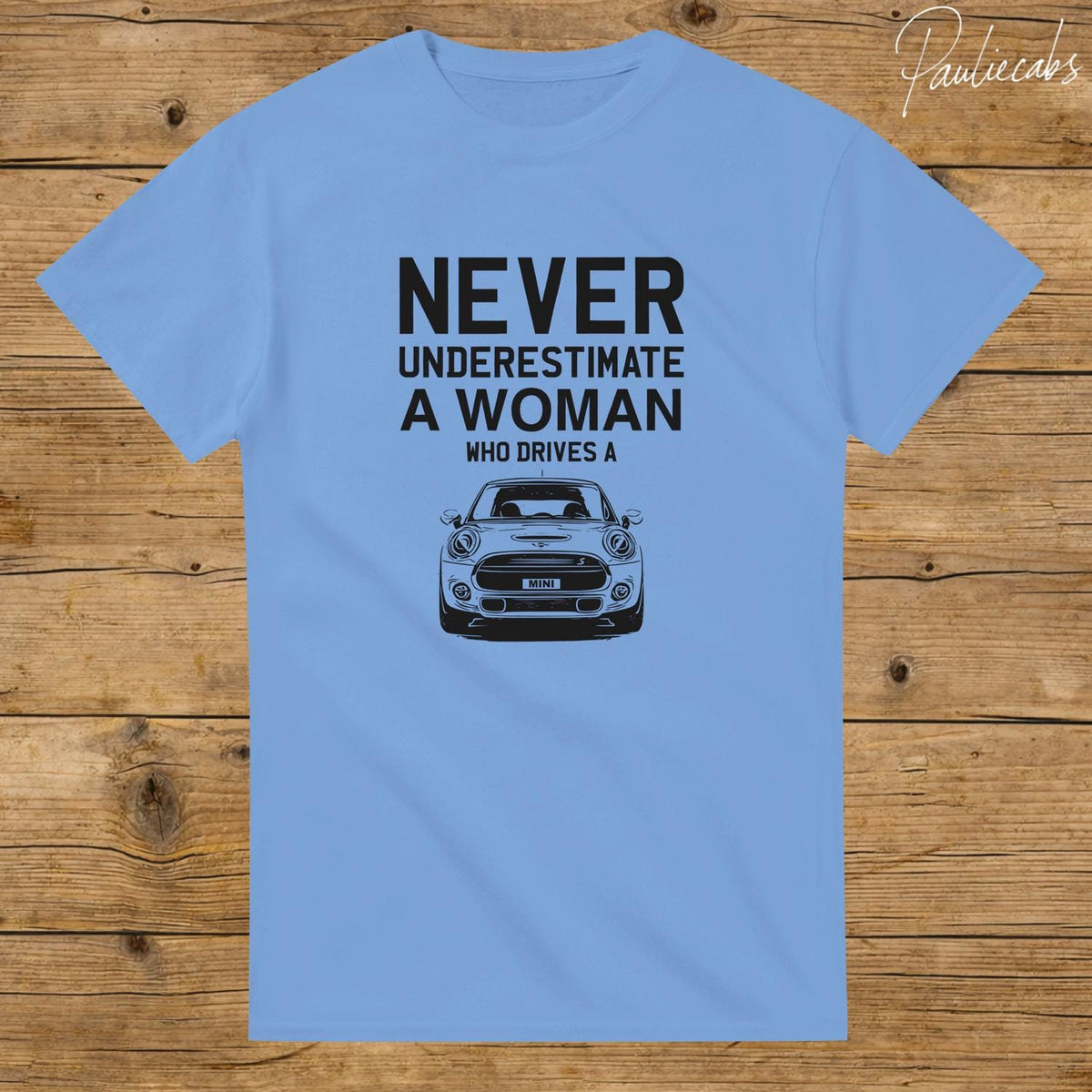 May include: A light blue short-sleeve t-shirt featuring a black graphic of a Mini Cooper car and the text: NEVER UNDERESTIMATE A WOMAN WHO DRIVES A. The design is centered on the chest of the shirt, which has a crew neckline and a standard fit.