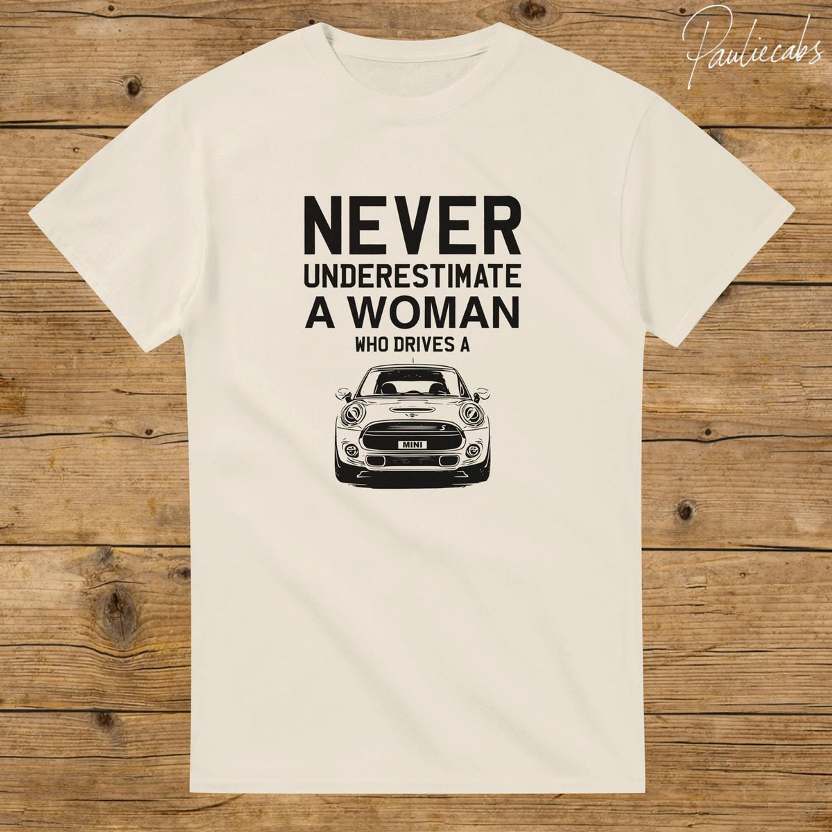 May include: A cream-colored short-sleeve t-shirt featuring black text that reads NEVER UNDERESTIMATE A WOMAN WHO DRIVES A, positioned above a black and white line art illustration of the front view of a Mini Cooper car with the word MINI on the plate.