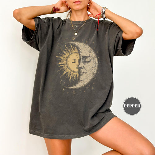 May include: Dark gray oversized t-shirt featuring a celestial design with a sun and moon face. The graphic is in shades of gold and beige, with stars surrounding the faces. The shirt has short sleeves and a relaxed fit.