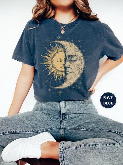May include: Navy blue t-shirt featuring a celestial design of a sun and moon embracing. The sun is depicted with golden rays, while the moon has a detailed, textured appearance. The shirt has a relaxed fit and is paired with blue jeans.