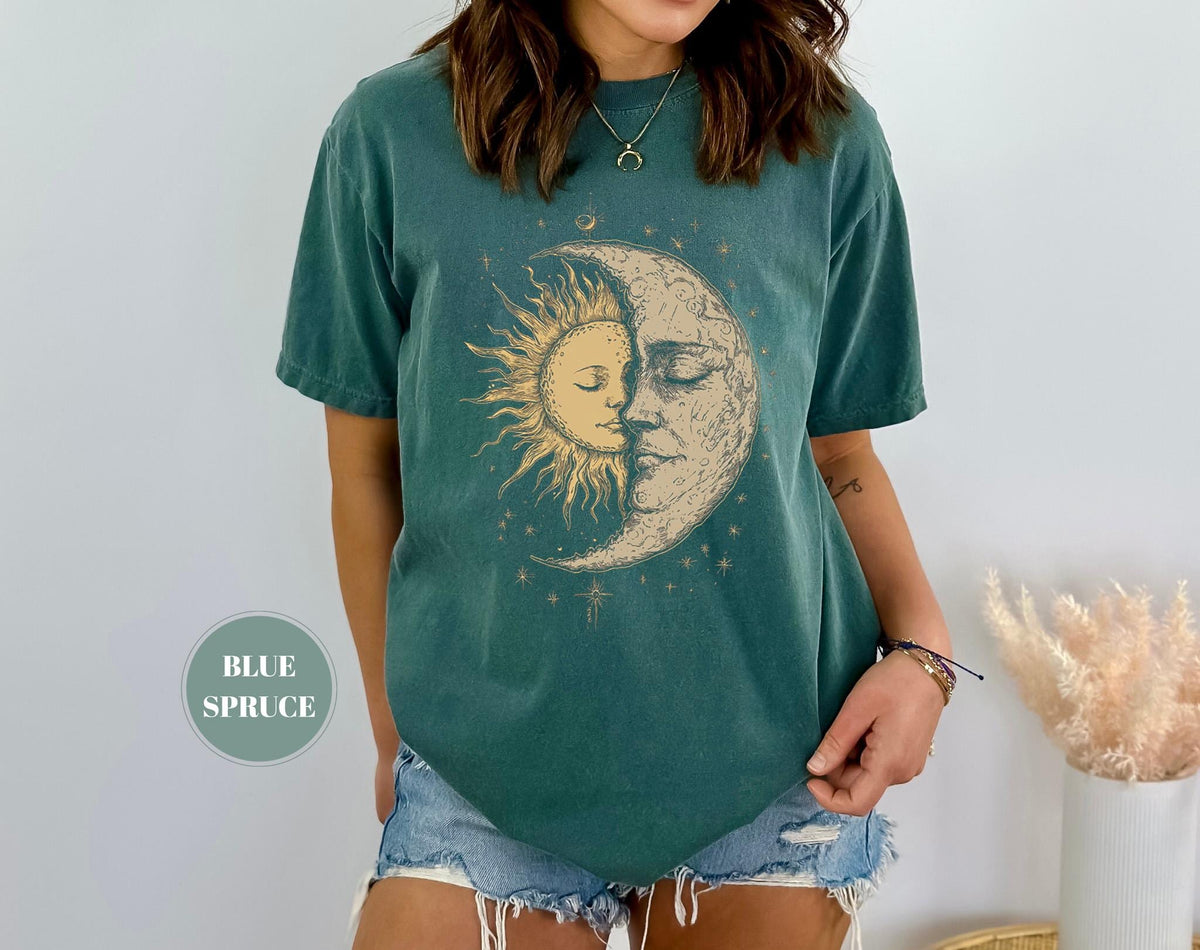 May include: A teal-colored t-shirt featuring a celestial design of a sun and moon embracing. The sun is golden, and the moon is cream-colored, with stars and celestial accents. The shirt has a relaxed fit and short sleeves. The text "BLUE SPRUCE" is in a circle.
