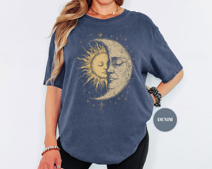May include: A navy blue t-shirt featuring a celestial design. The graphic depicts a sun and moon with faces, surrounded by stars. The shirt has short sleeves and a relaxed fit. The word "DENIM" is printed on a circular tag.