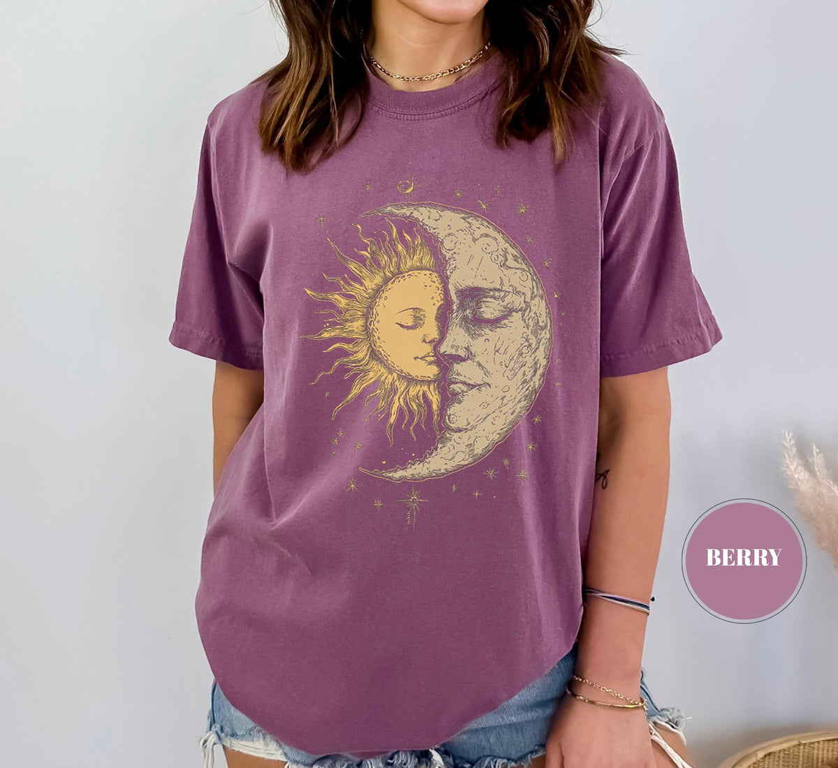 May include: Berry-colored t-shirt featuring a celestial design of a sun and moon embracing. The graphic is in shades of gold and cream, with the sun's rays radiating outward. The shirt has short sleeves and a relaxed fit.