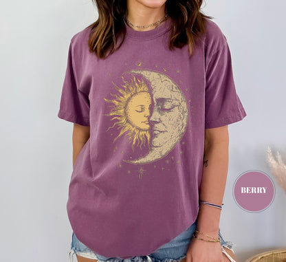 May include: Berry-colored t-shirt featuring a celestial design of a sun and moon embracing. The graphic is in shades of gold and cream, with the sun's rays radiating outward. The shirt has short sleeves and a relaxed fit.