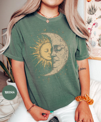 May include: A moss green t-shirt featuring a sun and moon graphic. The sun is depicted with radiating lines and a face, while the moon has a face with closed eyes. The shirt is a relaxed fit.