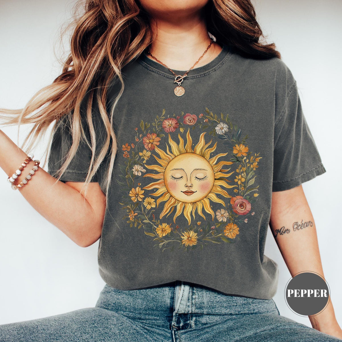 May include: Dark grey t-shirt featuring a sun graphic with closed eyes, surrounded by a floral wreath. The sun is yellow and orange, and the flowers are various colors. The shirt has a relaxed fit.