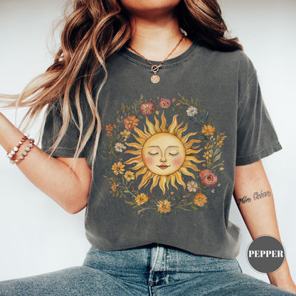 May include: Dark grey t-shirt featuring a sun graphic with closed eyes, surrounded by a floral wreath. The sun is yellow and orange, and the flowers are various colors. The shirt has a relaxed fit.