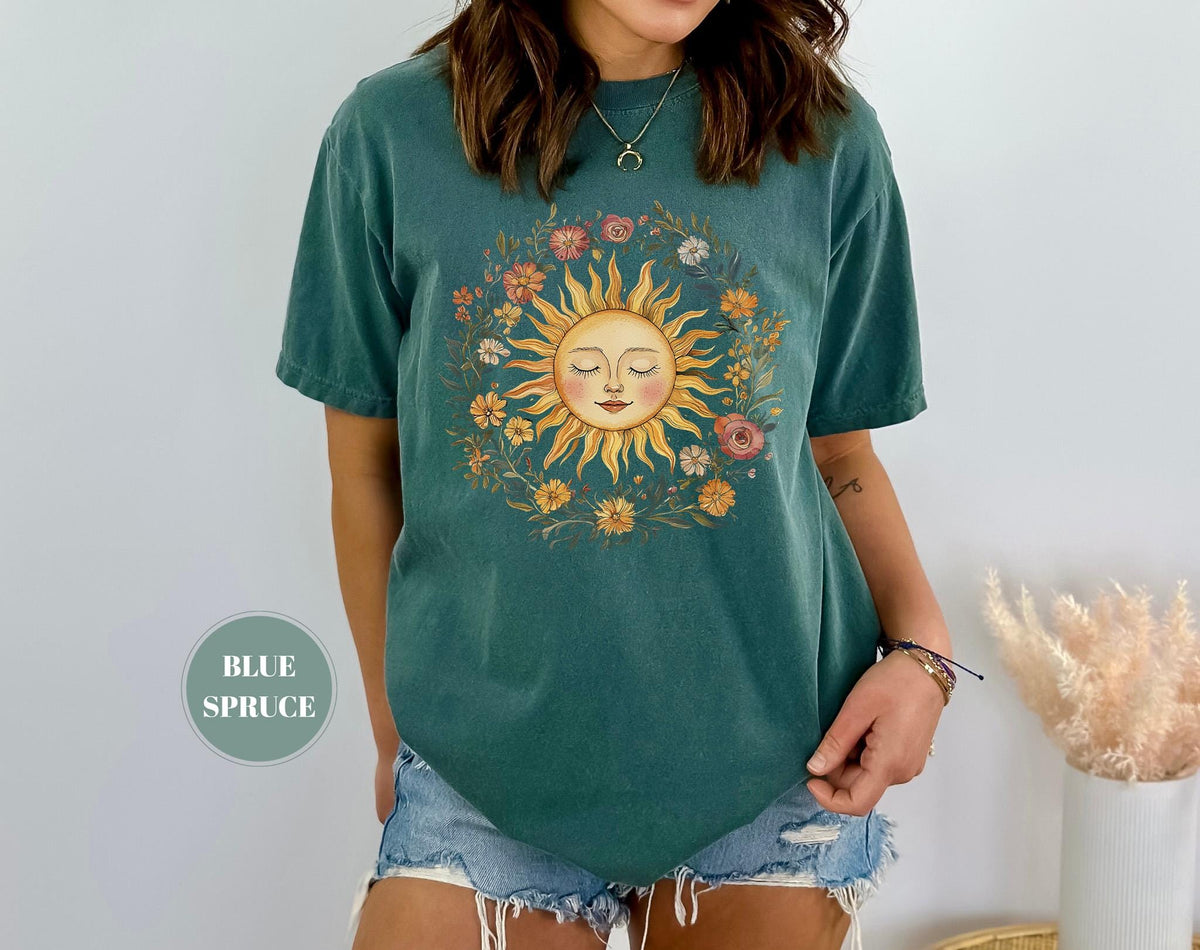 May include: A teal-colored t-shirt featuring a sun graphic surrounded by a floral wreath. The sun has a smiling face with closed eyes. The shirt is a short-sleeved, casual style. The text "BLUE SPRUCE" is in a circle on the bottom left.