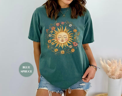 May include: A teal-colored t-shirt featuring a sun graphic surrounded by a floral wreath. The sun has a smiling face with closed eyes. The shirt is a short-sleeved, casual style. The text "BLUE SPRUCE" is in a circle on the bottom left.