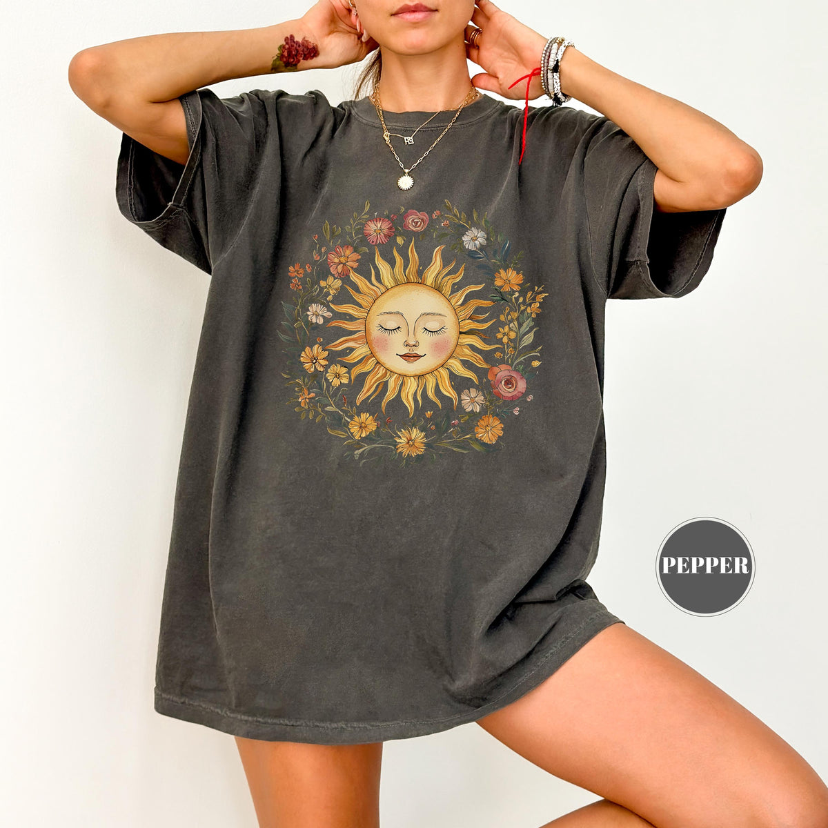 May include: Dark grey t-shirt featuring a smiling sun design surrounded by a floral wreath. The sun has closed eyes and a gentle expression. The shirt has short sleeves and a relaxed fit, perfect for casual wear.