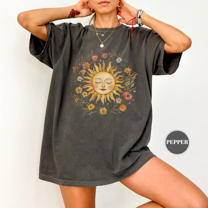 May include: Dark grey t-shirt featuring a smiling sun design surrounded by a floral wreath. The sun has closed eyes and a gentle expression. The shirt has short sleeves and a relaxed fit, perfect for casual wear.