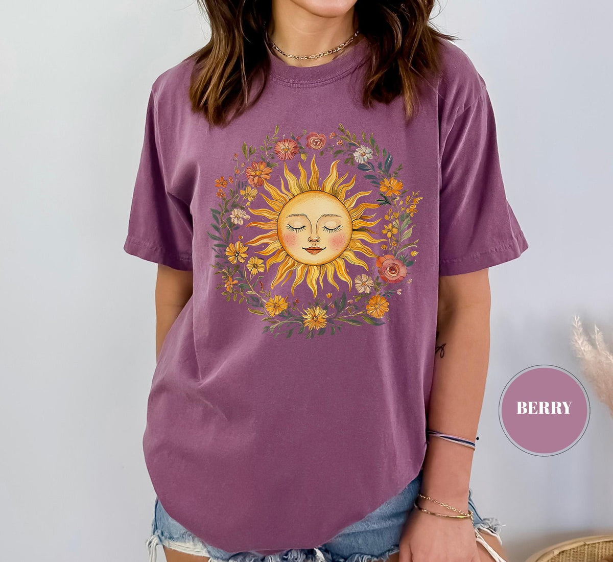 May include: Berry-colored t-shirt featuring a sun graphic with closed eyes, surrounded by a floral wreath. The sun has a smiling face and is in shades of yellow and orange. The flowers in the wreath are in various colors, including pink, yellow, and green.