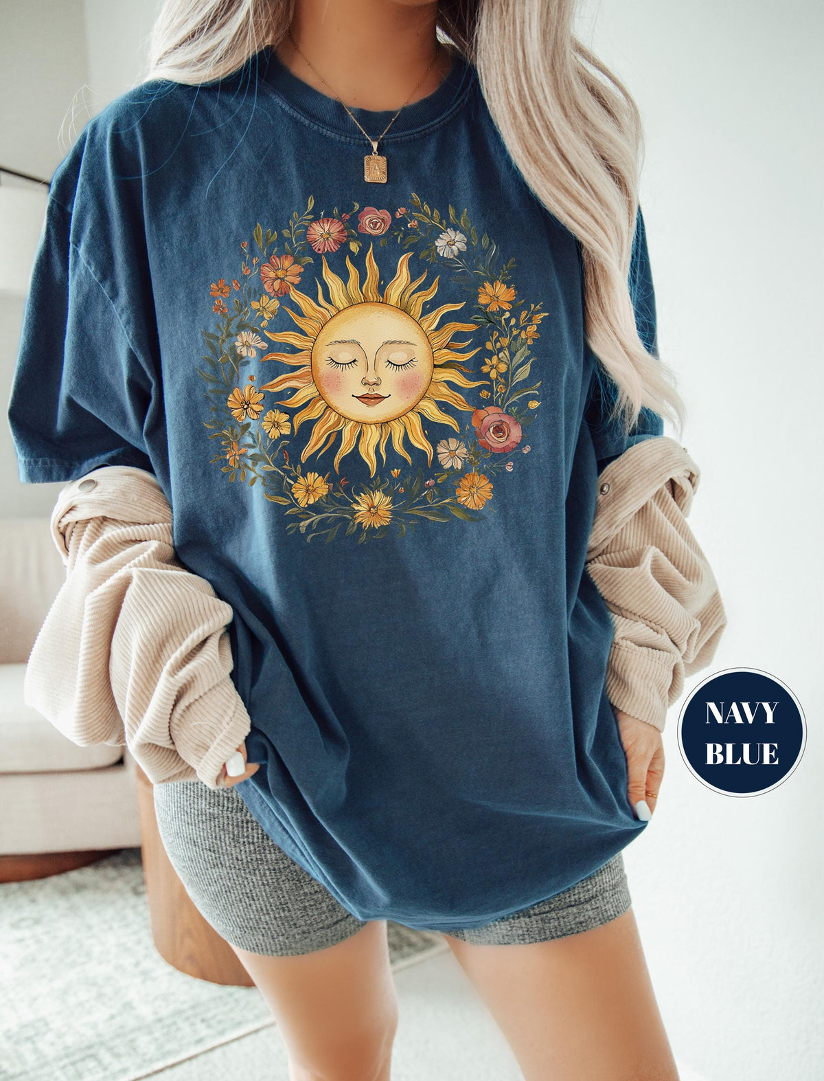 May include: Navy blue t-shirt featuring a sun graphic with closed eyes, surrounded by a floral wreath in shades of yellow, orange, pink, and green. The shirt has a relaxed fit and is labeled "NAVY BLUE" in a circular badge.
