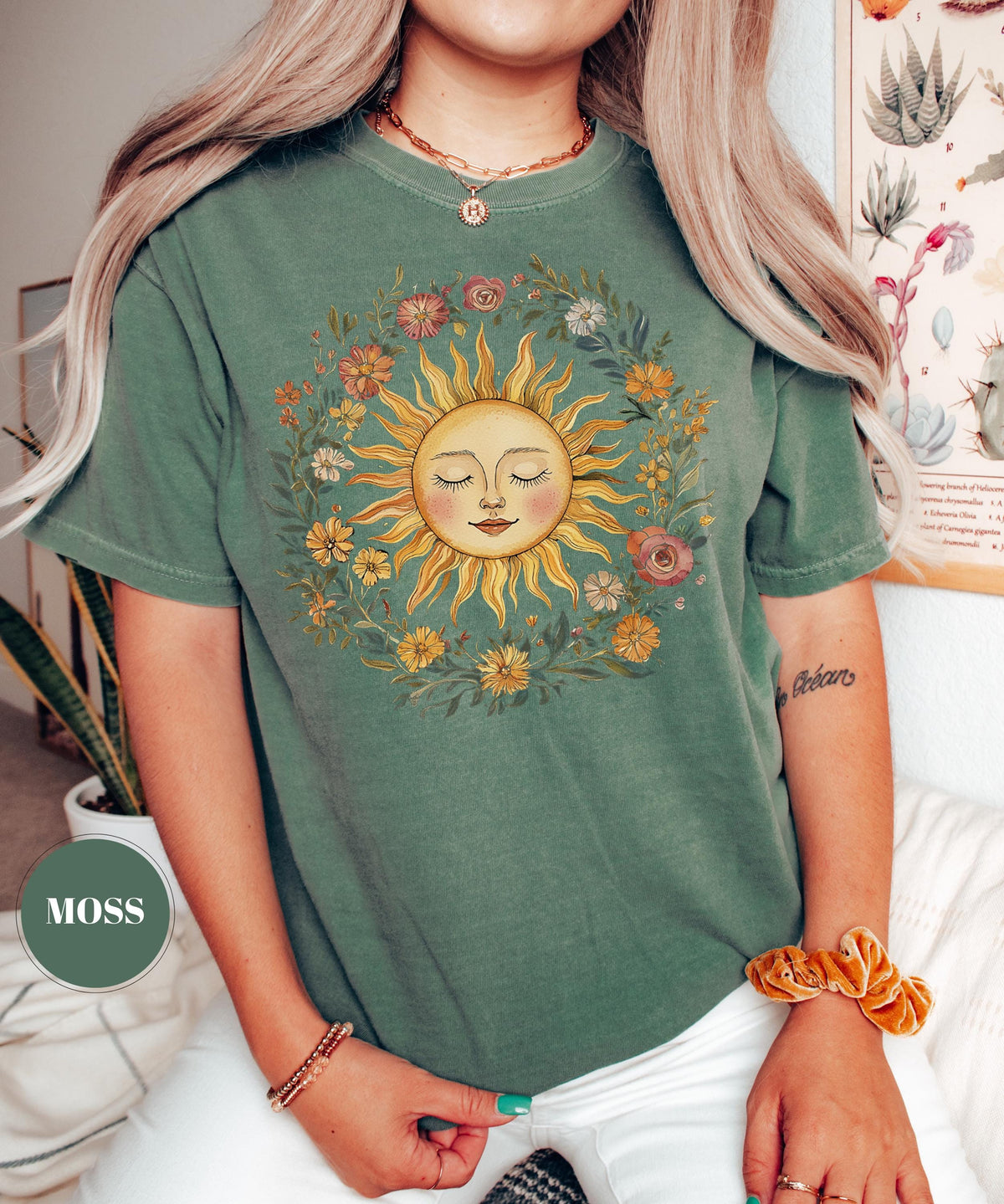 May include: Moss green t-shirt featuring a smiling sun graphic surrounded by a floral wreath. The sun has closed eyes and a warm yellow color. The shirt is a relaxed fit and has short sleeves.