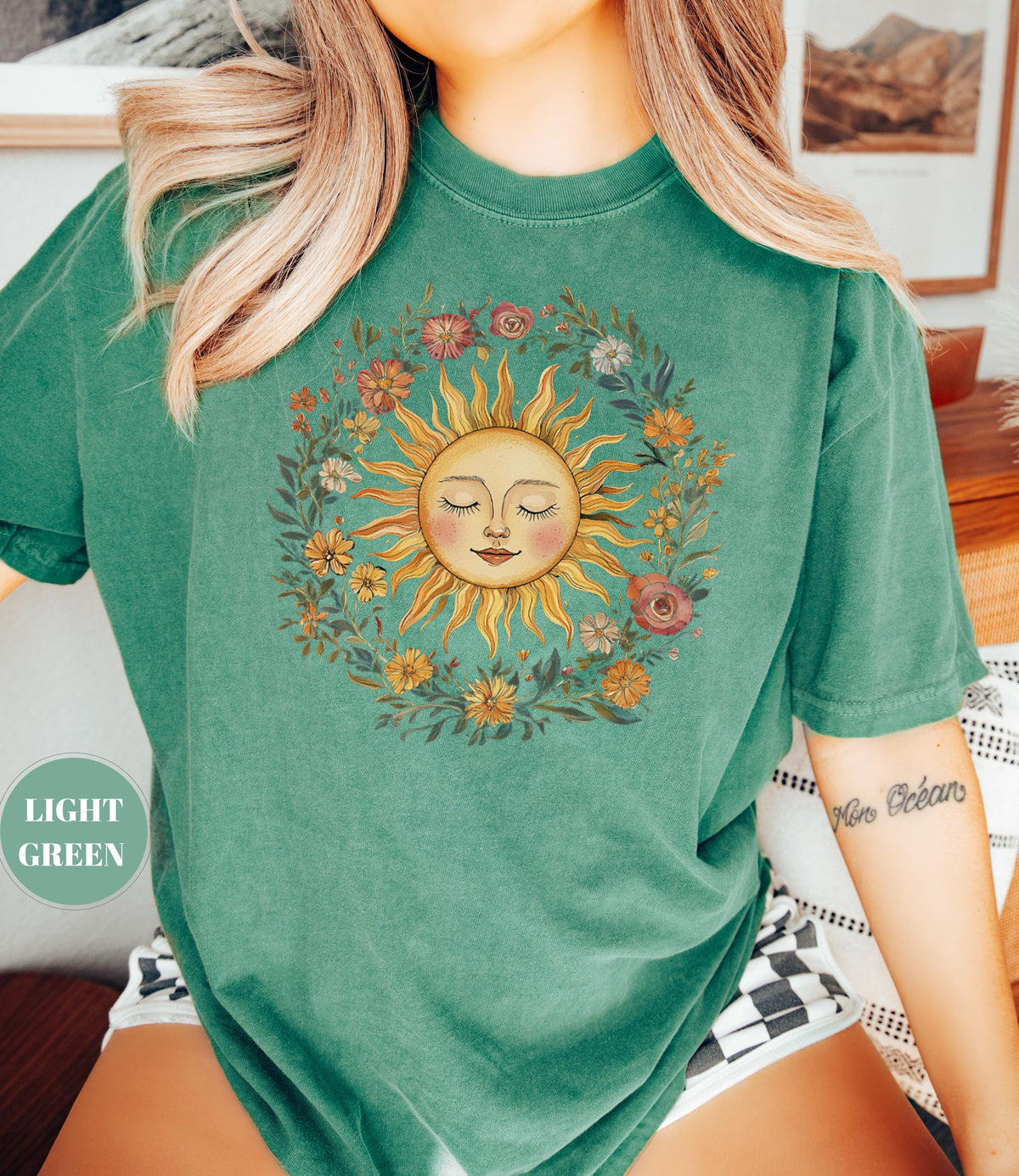 May include: Light green t-shirt featuring a sun graphic with a smiling face and closed eyes, surrounded by a floral wreath. The sun has yellow rays and a peach-colored face. The text "LIGHT GREEN" is on the left side of the shirt.