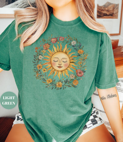 May include: Light green t-shirt featuring a sun graphic with a smiling face and closed eyes, surrounded by a floral wreath. The sun has yellow rays and a peach-colored face. The text "LIGHT GREEN" is on the left side of the shirt.