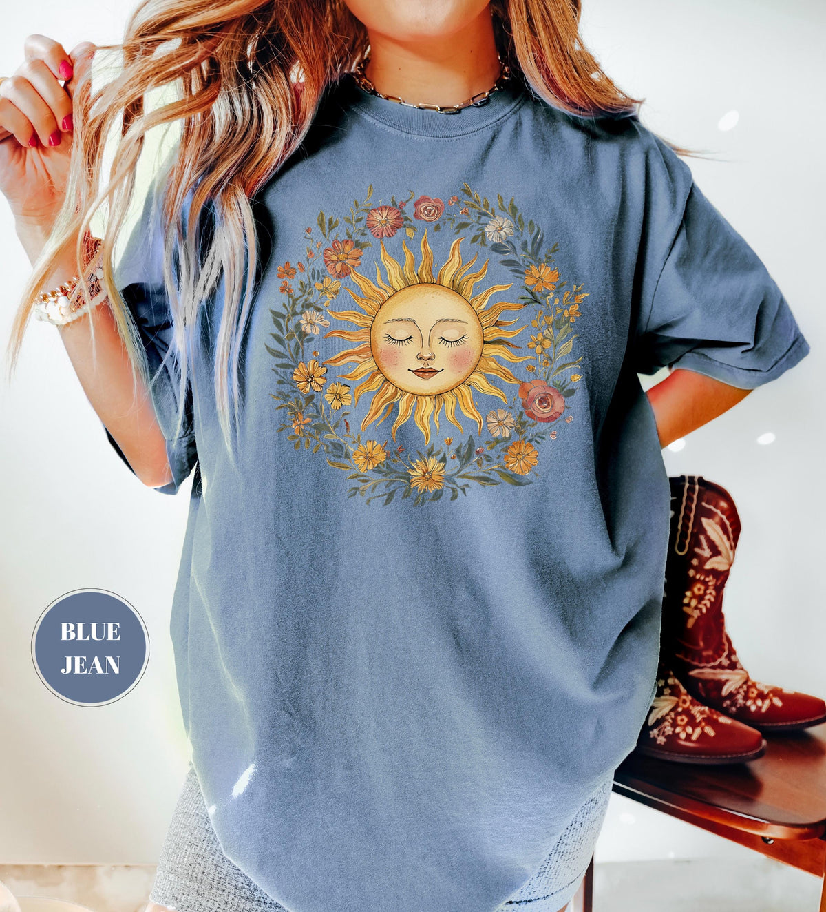 May include: Blue jean colored t-shirt featuring a smiling sun graphic surrounded by a floral wreath. The sun has closed eyes and a serene expression. The text "BLUE JEAN" is in a circle on the shirt.