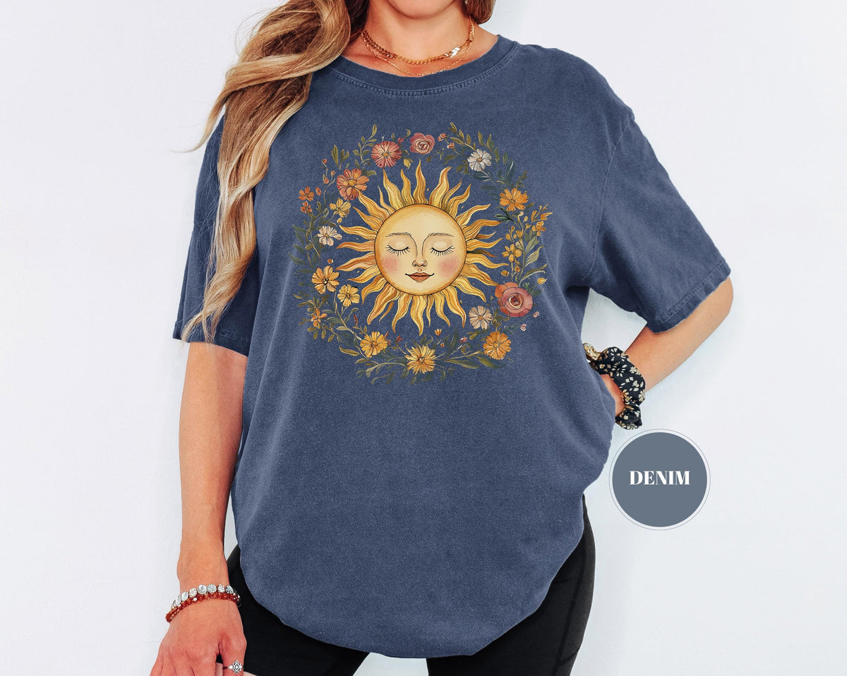May include: Denim blue t-shirt featuring a sun graphic with closed eyes, surrounded by a floral wreath. The sun has a smiling face and is in shades of yellow, orange, and peach. The flowers are in various colors, including pink and orange.