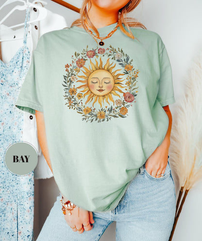 May include: A light green t-shirt featuring a sun design with a floral wreath. The sun has a smiling face and is surrounded by colorful flowers and leaves. The shirt is a relaxed fit.