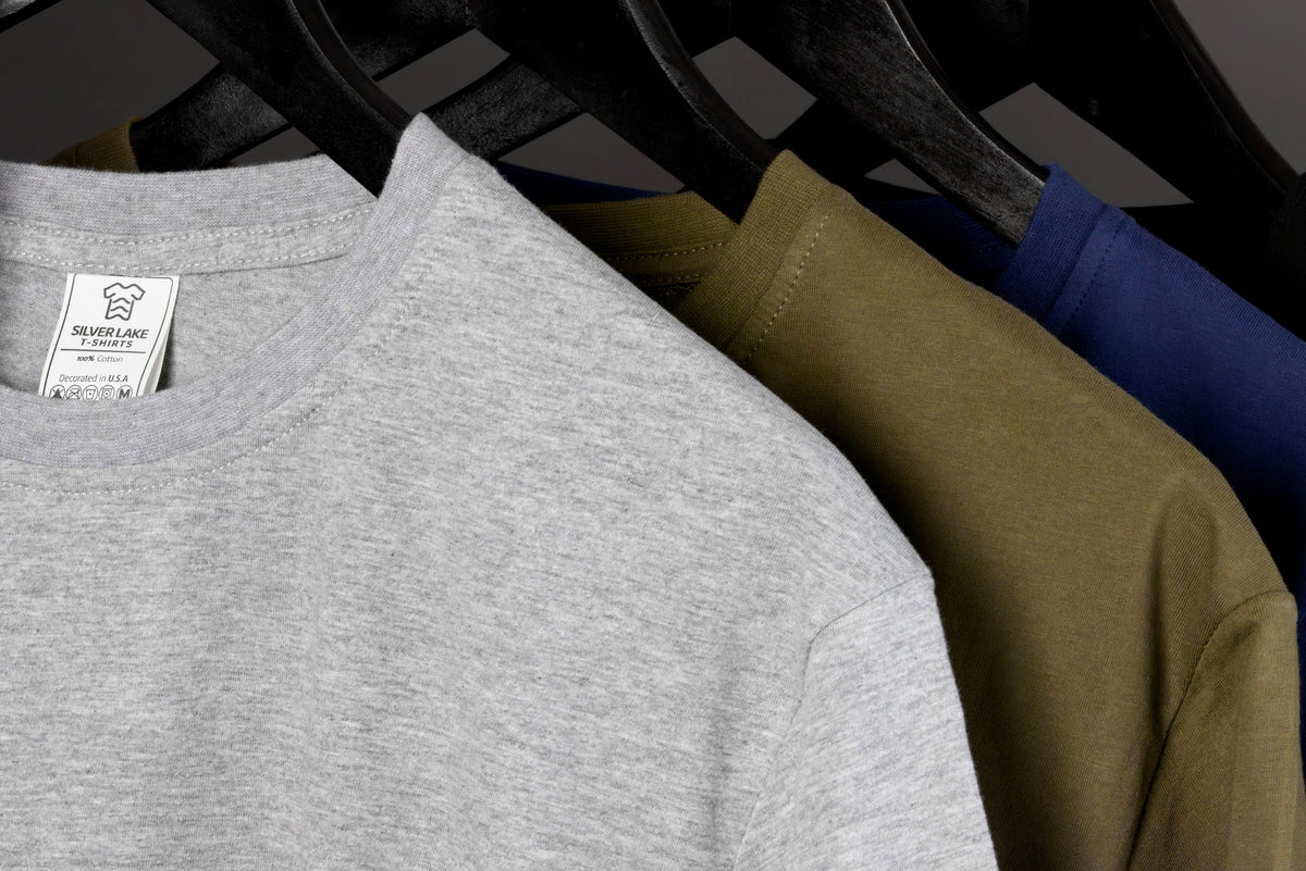 May include: Close-up of three t-shirts in gray, olive green, and navy blue hanging on a black hanger. The gray shirt has a tag that reads "Silver Lake T-Shirts" and "Designed in USA". The shirts are made of a soft material.