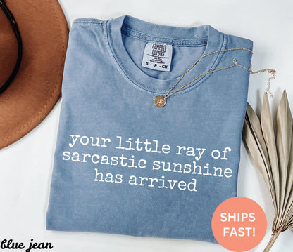 May include: A blue jean Comfort Colors t-shirt with the text "your little ray of sarcastic sunshine has arrived" in white. A gold necklace with a pendant is on the shirt. A brown hat is in the background.