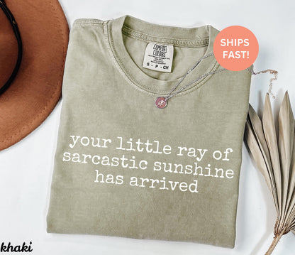 May include: Khaki-colored t-shirt with the phrase "your little ray of sarcastic sunshine has arrived" printed in white. A silver necklace with a pendant is on the shirt. The shirt has a Comfort Colors tag.