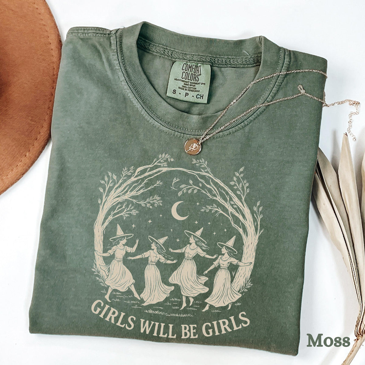 May include: A moss green short-sleeve t-shirt featuring a graphic of four figures in pointed hats dancing in a circle under a crescent moon and tree branches. The text "GIRLS WILL BE GIRLS" is printed below the graphic in a light beige color.