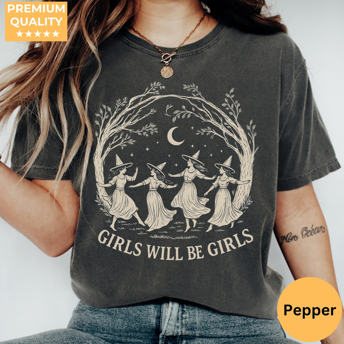 May include: A charcoal gray short-sleeve t-shirt featuring a cream-colored graphic of four figures in pointed hats dancing in a circle under a crescent moon and stars, framed by tree branches, with the text GIRLS WILL BE GIRLS printed below.