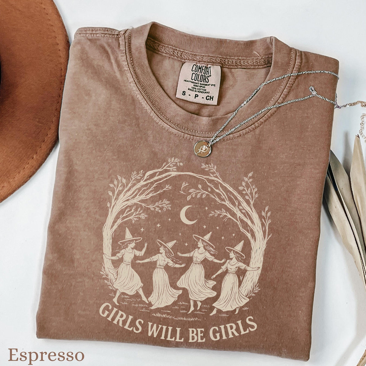 May include: An espresso-colored t-shirt featuring a graphic of four figures in witch hats dancing in a circle under a crescent moon and stars, framed by tree branches. The text "GIRLS WILL BE GIRLS" is printed below the illustration in a light color.