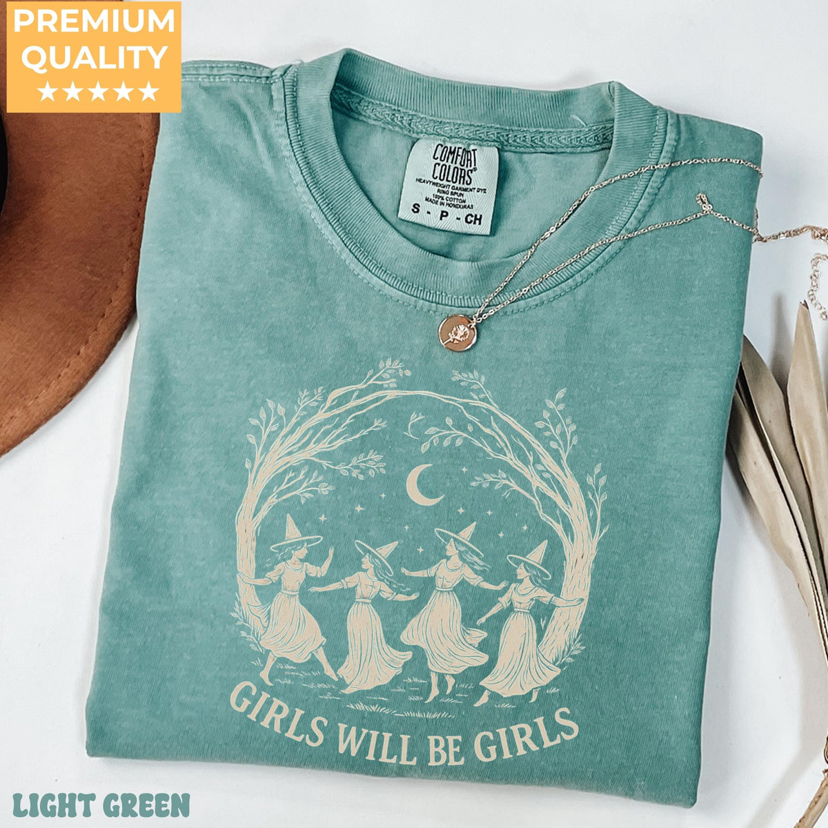 May include: A light green t-shirt featuring a graphic of four figures in pointed hats dancing under a crescent moon and tree branches. The text "GIRLS WILL BE GIRLS" is printed below the illustration. The shirt has a Comfort Colors brand label.