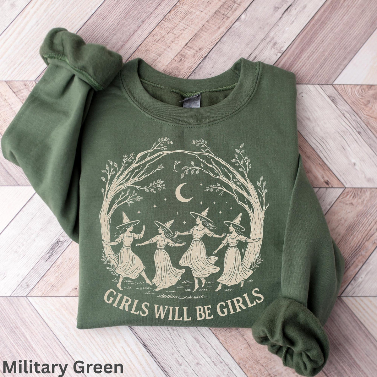 May include: A military green crewneck sweatshirt featuring a cream-colored graphic of four figures wearing pointed hats dancing in a circle under a crescent moon and tree branches, with the text "GIRLS WILL BE GIRLS" printed below the illustration.