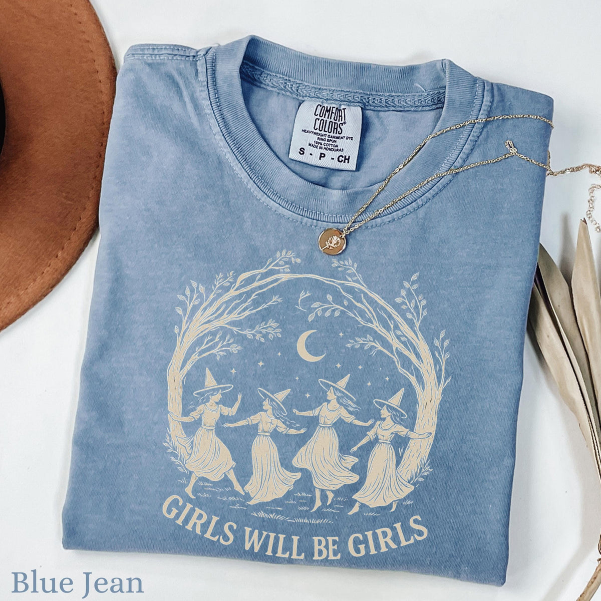 May include: A blue jean colored t-shirt with a cream-colored graphic print featuring four figures in pointed hats dancing in a circle under a crescent moon and tree branches, with the text "GIRLS WILL BE GIRLS" printed below the illustration.