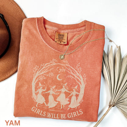 May include: A terracotta-colored short-sleeve t-shirt featuring a cream-colored graphic of four figures in witch hats dancing in a circle under a crescent moon and tree branches, with the text "GIRLS WILL BE GIRLS" printed below the graphic.