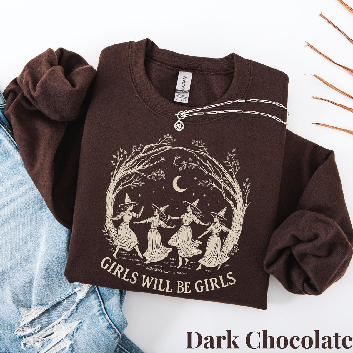 May include: A dark chocolate brown crewneck sweatshirt featuring a cream-colored graphic of four figures in pointed hats dancing in a circle under a crescent moon and tree branches, with the text "GIRLS WILL BE GIRLS" printed below the illustration.