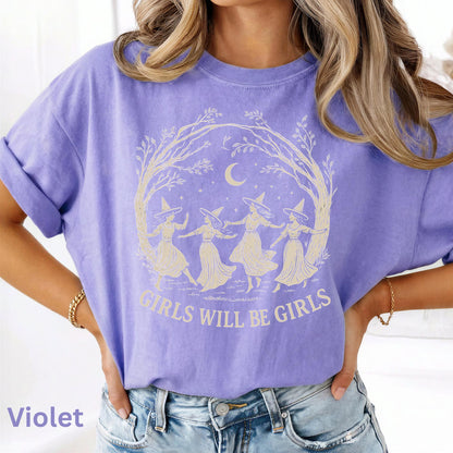 May include: A violet short-sleeved t-shirt featuring a cream-colored graphic of four figures wearing pointed hats dancing in a circle under a crescent moon and tree branches, with the text "GIRLS WILL BE GIRLS" printed in a bold font below.