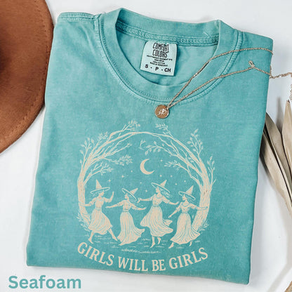 May include: A seafoam green short-sleeve t-shirt with a graphic print of four figures wearing pointed hats dancing under a crescent moon and tree branches. The text "GIRLS WILL BE GIRLS" is printed below the illustration in a cream-colored font.