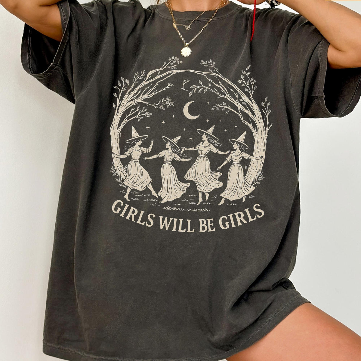 May include: A charcoal gray short-sleeve t-shirt featuring a beige graphic print of four figures in witch hats dancing under a crescent moon and tree branches. The text "GIRLS WILL BE GIRLS" is printed in a beige arched font below the illustration.