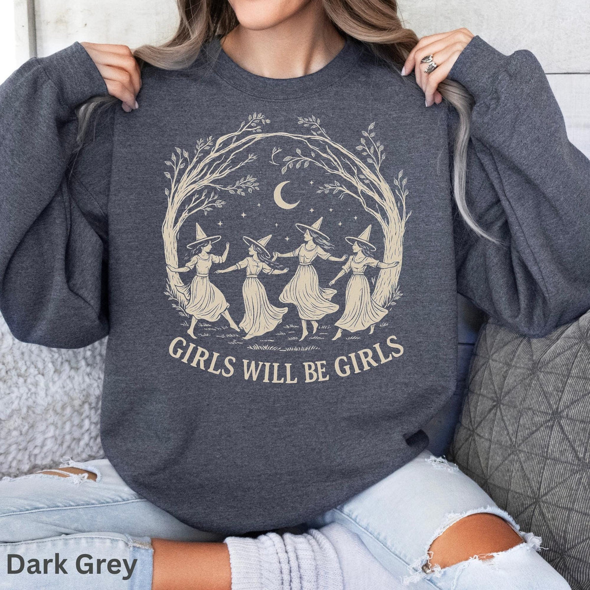 May include: A dark grey crewneck sweatshirt featuring a light-colored graphic of four figures wearing pointed hats dancing in a circle under a crescent moon and tree branches, with the text GIRLS WILL BE GIRLS printed below the illustration.
