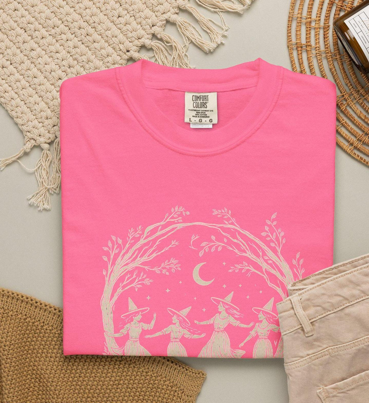 May include: A bright pink t-shirt featuring a cream-colored graphic print of four figures wearing pointed hats dancing under a crescent moon and tree branches. The shirt has a crew neckline and a Comfort Colors brand label at the inner neck.