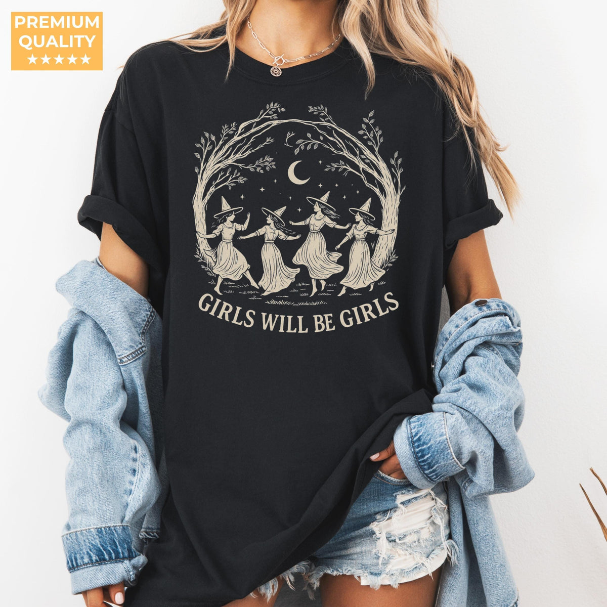 May include: A black short-sleeve t-shirt featuring a cream-colored graphic of four figures wearing pointed hats dancing in a circle under a crescent moon and tree branches, with the text GIRLS WILL BE GIRLS printed in a bold serif font below.