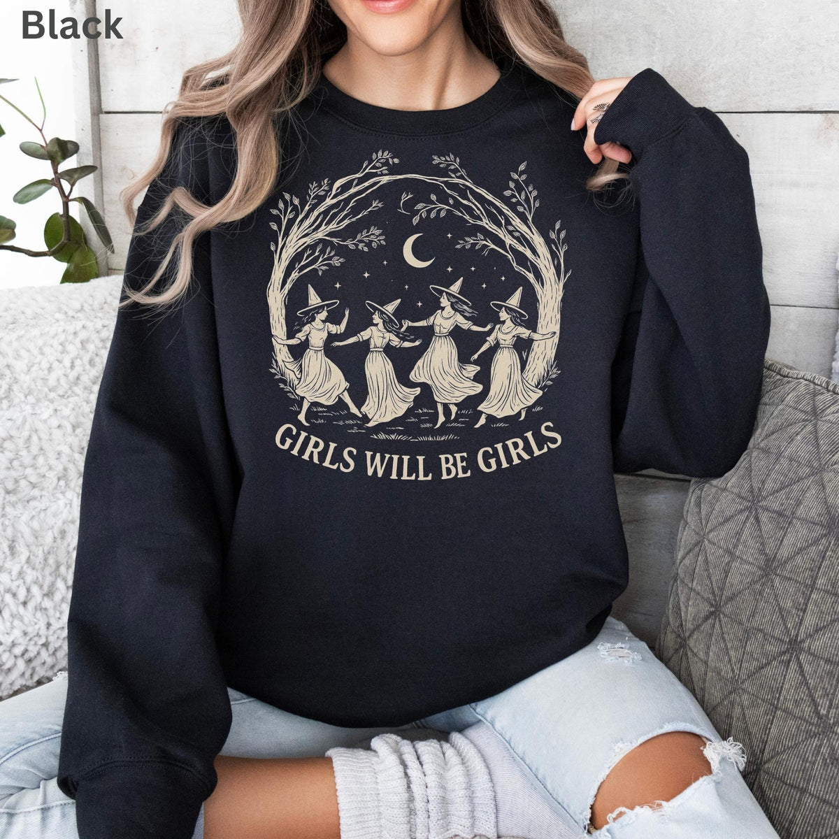 May include: A black long-sleeve sweatshirt featuring a beige graphic print of four figures in pointed hats dancing in a circle under a crescent moon and tree branches, with the text "GIRLS WILL BE GIRLS" printed in a curved font below the illustration.