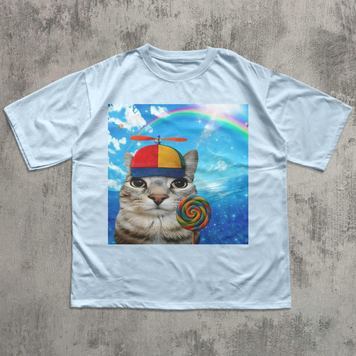 May include: Light blue t-shirt featuring a whimsical graphic of a cat wearing a propeller hat and holding a colorful lollipop. The background includes a rainbow and a blue sky with clouds. The shirt is made of a soft material.
