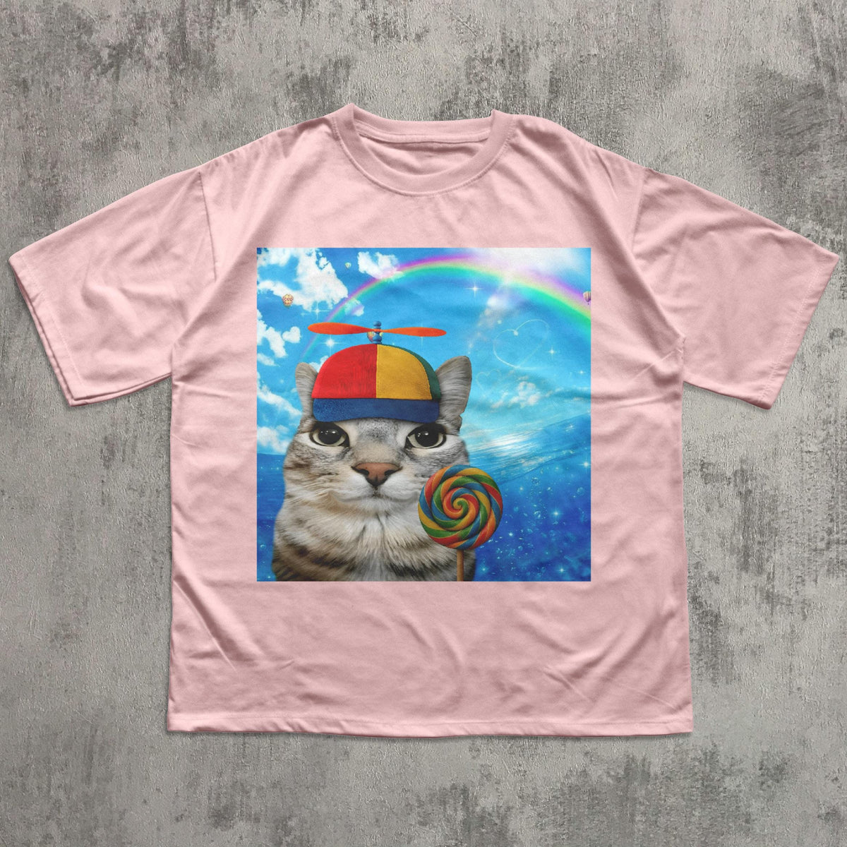 May include: A light pink t-shirt featuring a graphic of a cat wearing a colorful hat and holding a lollipop. The graphic includes a rainbow and a blue sky background. The cat has a striped pattern.
