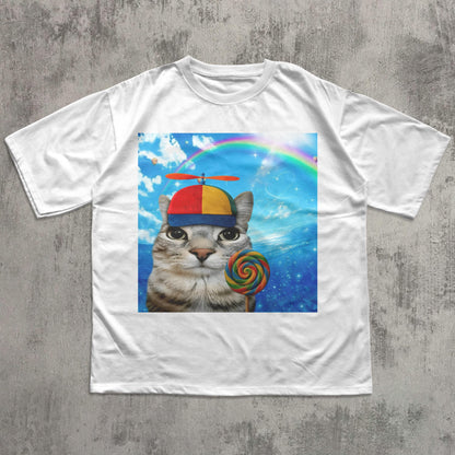 May include: White t-shirt featuring a whimsical graphic of a cat wearing a colorful hat with a propeller and holding a lollipop. The cat is set against a backdrop of a rainbow and a blue sky with clouds. A fun and playful design.