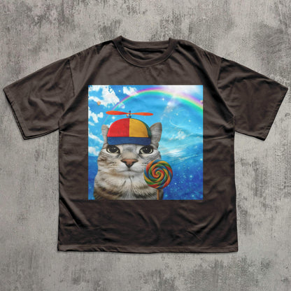 May include: Brown t-shirt featuring a whimsical graphic of a cat wearing a colorful propeller hat and holding a lollipop. The graphic includes a rainbow and a blue sky background. The shirt is made of a soft material.