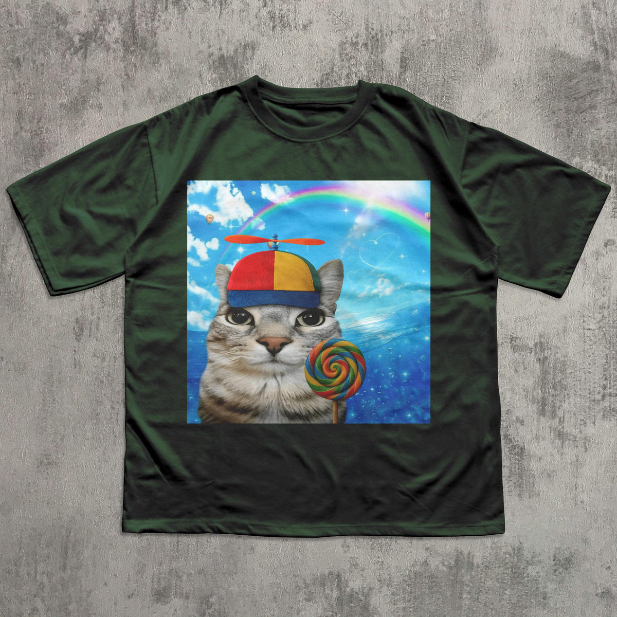 May include: Dark green t-shirt featuring a graphic of a cat wearing a propeller hat and holding a lollipop. The graphic includes a rainbow and a blue sky background. The cat has a striped pattern.