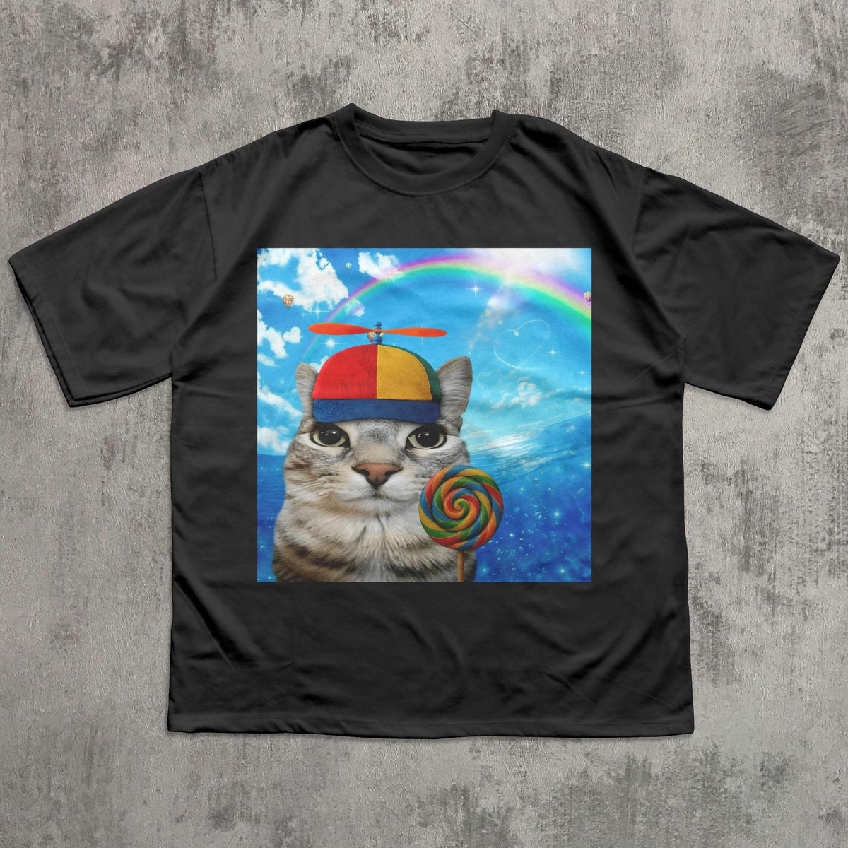 May include: Black t-shirt featuring a whimsical graphic of a cat wearing a propeller hat and holding a colorful lollipop. The background includes a rainbow and a blue sky with clouds. The cat's expression is curious.