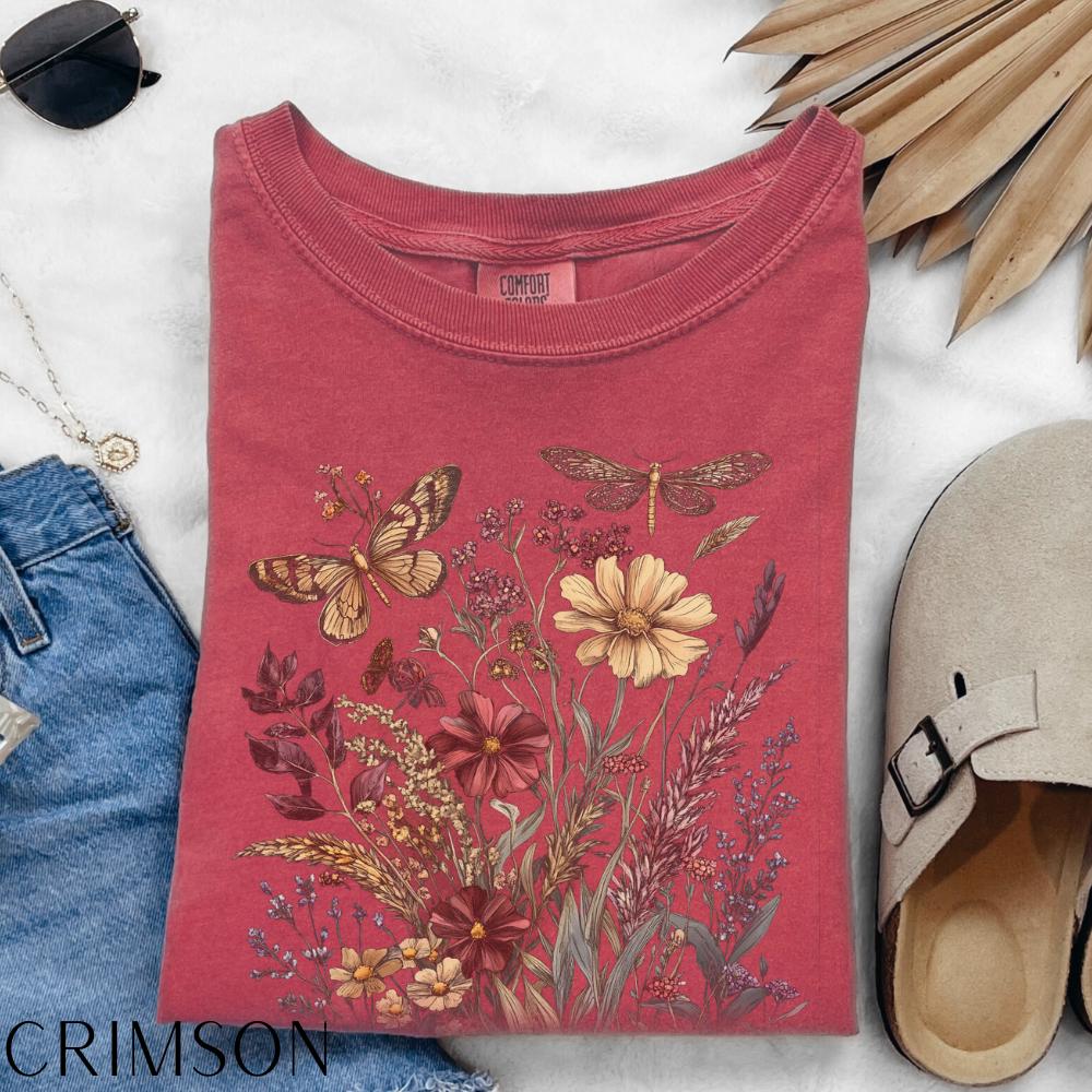 May include: Crimson-colored t-shirt featuring a floral and insect design. The graphic includes a butterfly, dragonfly, and various flowers in shades of yellow, red, and brown. The shirt is folded, with the word "CRIMSON" visible at the bottom.